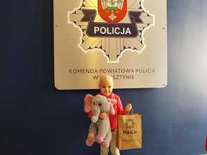 roksana stoi na tle logo wolsztynskiej policji, a w reku trzyma manskkotke wielkopolskiej policji i paczke z prezentami