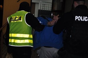 na zdjęciu zatrzymany poszukiwany mężczyzna z kajdankami na rękach prowadzony przez dwóch policjantów - zdjęcie zrobione z tyłu