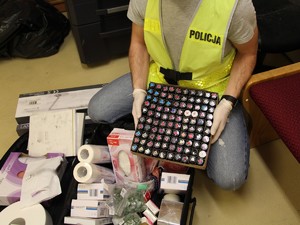 Na zdjęciu kucający policjant w kamizelce z napisem &quot;Policja&quot;. Na rękach ma gumowe rękawiczki. Trzyma karton z lakierami do paznokci.