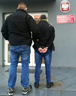 Nieumundurowany policjant trzyma zatrzymanego, skutego w kajdankach. Oboje stoją tyłem przed wejściem do komendy.