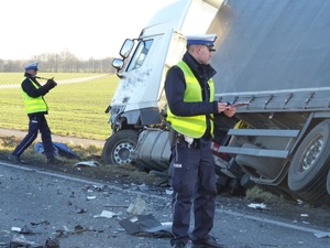 dwóch funkcjonariuszy sporządzających notatkę i zdjęcia z miejsca wypadku, a w tle widok na pobocze i przechylonego bokiem samochodu ciężarowego