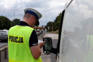 Policjant ruchu drogowego sprawdza dokumenty zatrzymanego kierowcy białego busa.