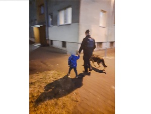 Policjant z psem służbowym idzie i trzyma za rękę dziecko - zdjęcie jest zrobione na z tyłu, niewyraźne, bo osoby się poruszają.