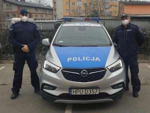 Dwóch policjantów umundurowanych stoi po obu stronach oznakowanego radiowozu - pozują do zdjęcia w maseczkach higienicznych na twarzach.
