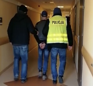 Policjanci w ubraniach cywilnych - jeden w kamizelce z napisem POLICJA prowadzą zatrzymanego mężczyznę.
