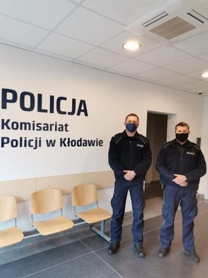 Dwóch umundurowanych policjantów stoi obok siebie w maseczkach, przy duym logo umieszczonym na cianie - Komisariat Policji w Kodawie