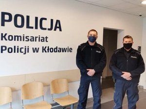 Dwóch umundurowanych policjantów stoi obok siebie w maseczkach, przy dużym logo umieszczonym na ścianie - Komisariat Policji w Kłodawie