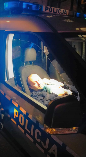 Zdjęcia zrobione na zewnątrz - ukazuje wnętrze radiowozu, w którym siedzi policjant, a na ramieniu trzyma śpiącego niemowlaka.