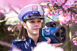 Policjantka trzyma radar schowana w drzewie z kwiatami koloru różowego