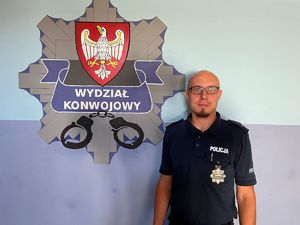 Policjant mł. asp. Szymon Karolewicz w mundurze na tle logo Wydziału Konwojowego KWP w Poznaniu