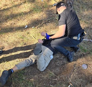 policjant zakuwa zatrzymanego, który leży na trawie w kajdanki; policjant z zatrzymanym idą przez las