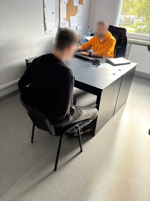 policjant siedzi przy biurku, na blacie dokumenty. naprzeciwko siedzi zatrzymany z rękoma skutymi kajdankami z tyłu