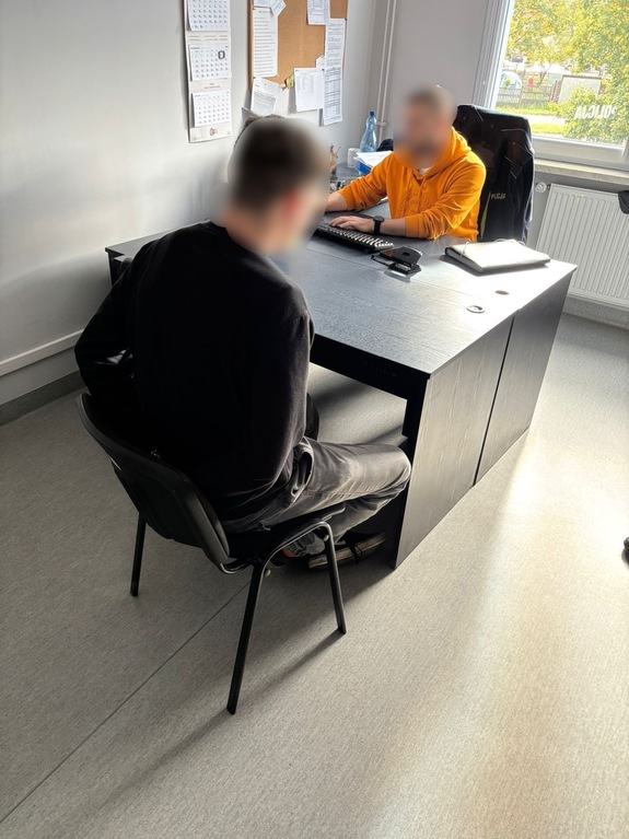policjant siedzi przy biurku, na blacie dokumenty. naprzeciwko siedzi zatrzymany z rękoma skutymi kajdankami z tyłu