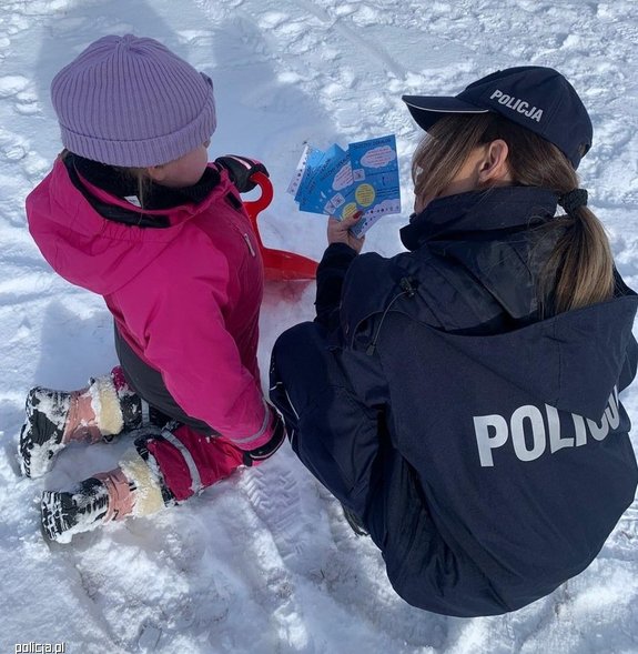 zima, policjantka kuca przy dziewczynce, pokazuje jej ulotki dotyczące bezpieczeństwa w zimie
